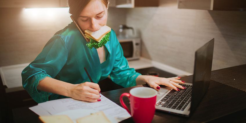 Woman eating a sandwich while writing and typing on a computer.