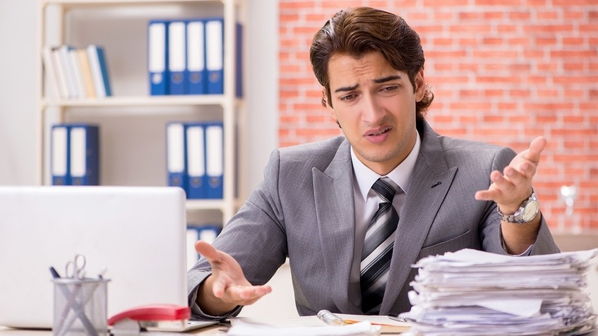Man in a business suit looking frustrated at a pile of papers on his desk.